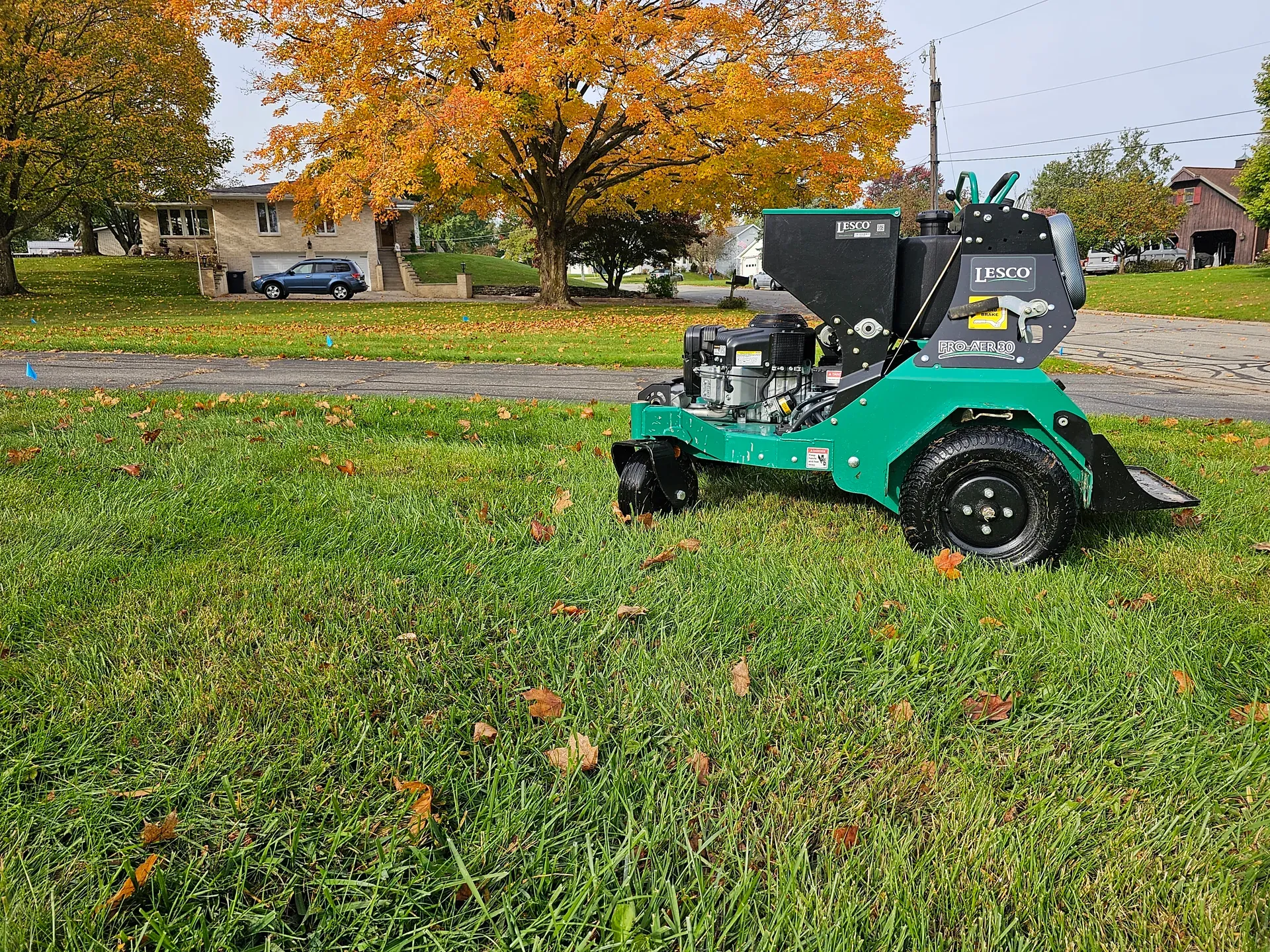 A GreenWorks technician running a core aerator across a green lawn ringed with fall foliage