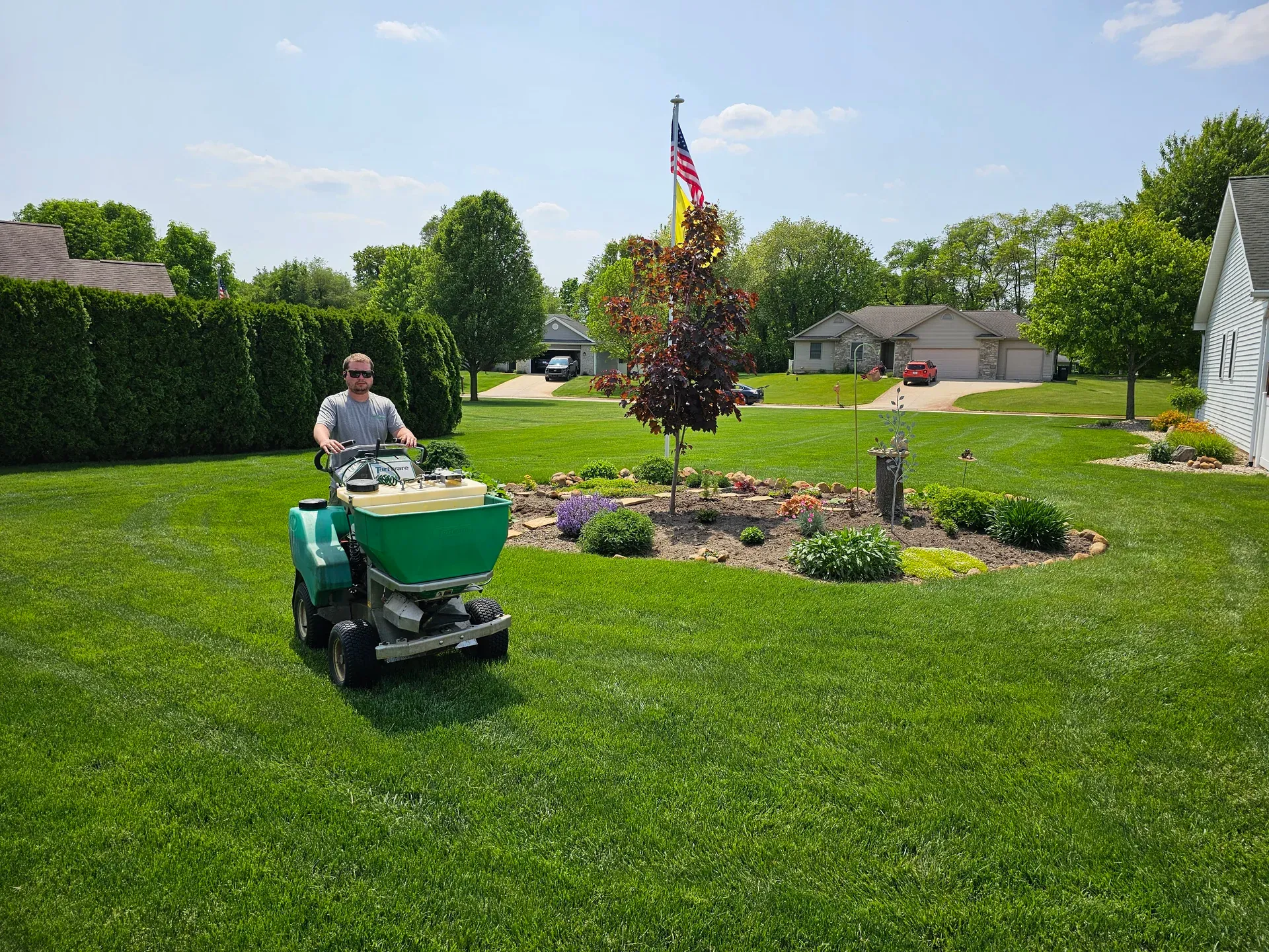 A GreenWorks technician driving a ride-on spreader past a flagpole and landscaped bed